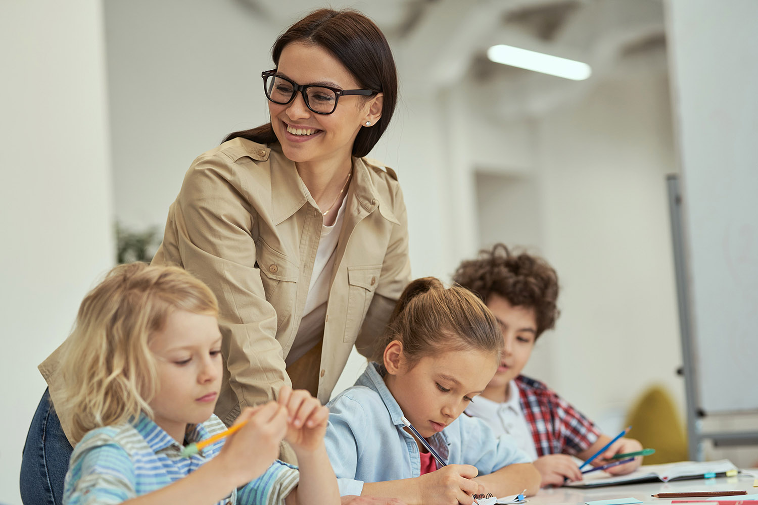 Glückliche junge Lehrerin mit Brille, die ihren kleinen Schülern in einem Klassenzimmer hilft. Kinder sitzen am Tisch und lernen in der Grundschule. Bildungskonzept für Kinder. Schulbegleitung bekommen MDLI
